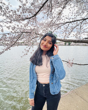 Samidha Sane stands in front of a cherry blossom tree and a body of water.