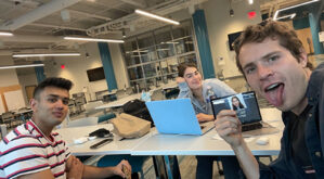 Shivam sits at a table with two others. Student intern Annie sits in the middle with her head in her hand. Alex sticks his tongue out and holds up a tablet.
