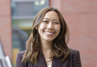 A headshot of a person smiling, wearing a blazer, with a blurred building background.