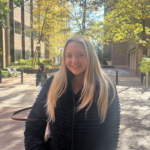 A person smiling outdoors on a sunlit brick pathway, with trees and a building in the background.