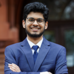 A person wearing glasses and a blue suit with a tie, smiling and standing with arms crossed in front of a building.