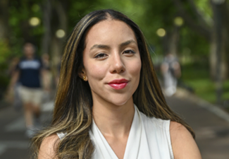 Headshot of a person with long hair wearing a sleeveless white top, standing on a path in a park with trees and people in the background.