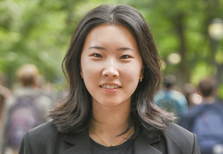 A headshot of a person with shoulder-length hair, wearing a black top and jacket, standing outdoors with a blurred background of trees and people.