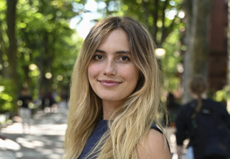 A headshot of a person with long hair, smiling outdoors on a tree-lined pathway. The background is slightly blurred with people walking.