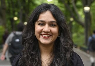 A person with long, dark hair is smiling outdoors with a blurred background of trees and a pathway.