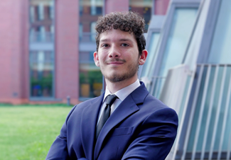 chance headshot A headshot of a person in a suit and tie, standing outdoors with a building and glass panels in the background.
