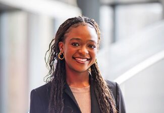 ebun headshot Headshot of a smiling person with long braided hair, wearing a black suit jacket and a light-colored top, standing indoors with blurred background.