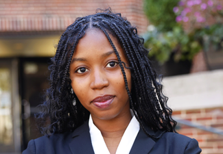 malaika headshot Headshot of a person with braided hair in a black blazer and white shirt, standing outside with a blurred background of a brick building and flowers.