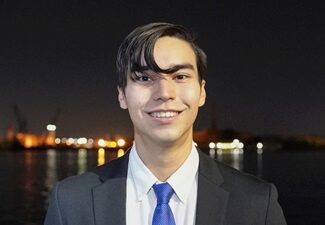 matthew headshot Headshot of a person in a suit and blue tie, smiling against a night-time cityscape with water and lights in the background.