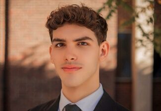 tareq headshot A headshot of a person with short curly hair, wearing a suit and tie, standing outdoors with a brick wall in the background.
