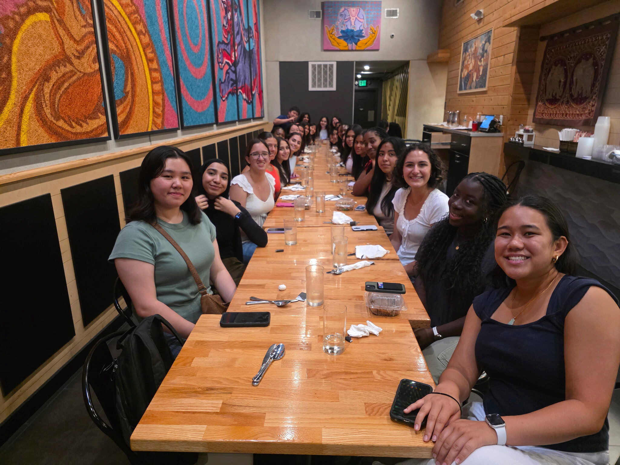 3484018134464184723 A large group of people sitting at a long dining table in a restaurant, smiling at the camera. The walls feature colorful artwork.
