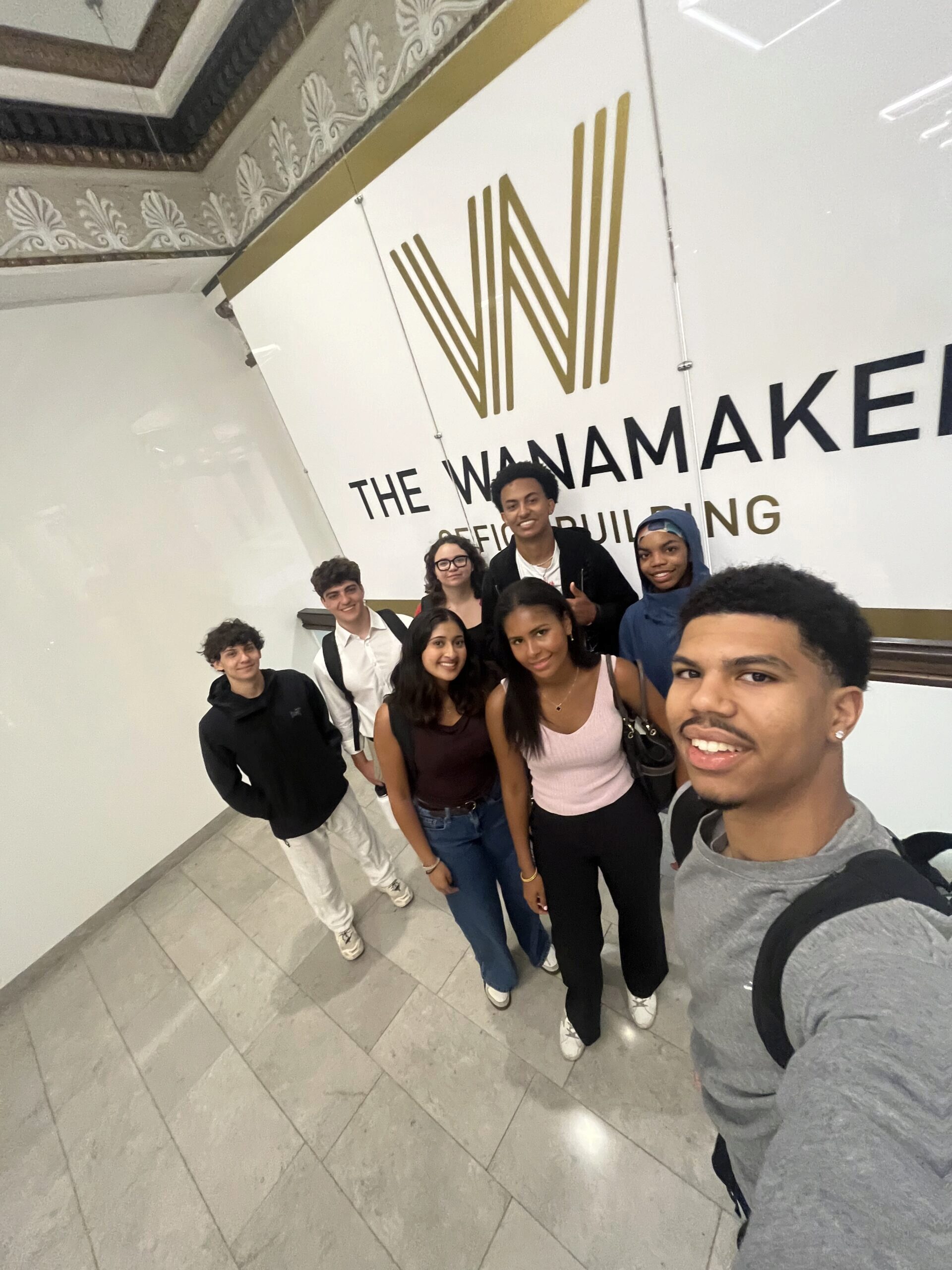IMG_3619 A group of people smiling and posing for a selfie in front of a sign that reads "The Wanamaker Office Building." They are standing on a tiled floor with ornate ceiling decorations visible.