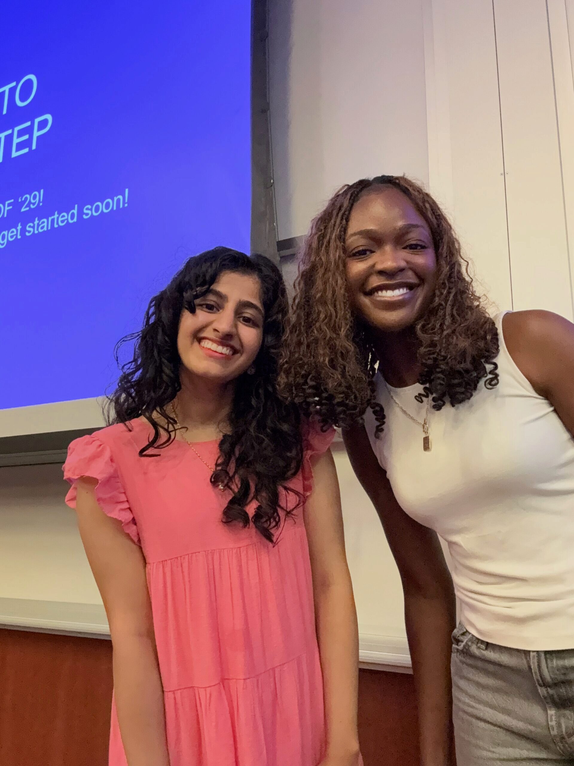 IMG_6661 Two people posing and smiling in front of a blue presentation screen, one wearing a pink dress and the other in a white sleeveless top.
