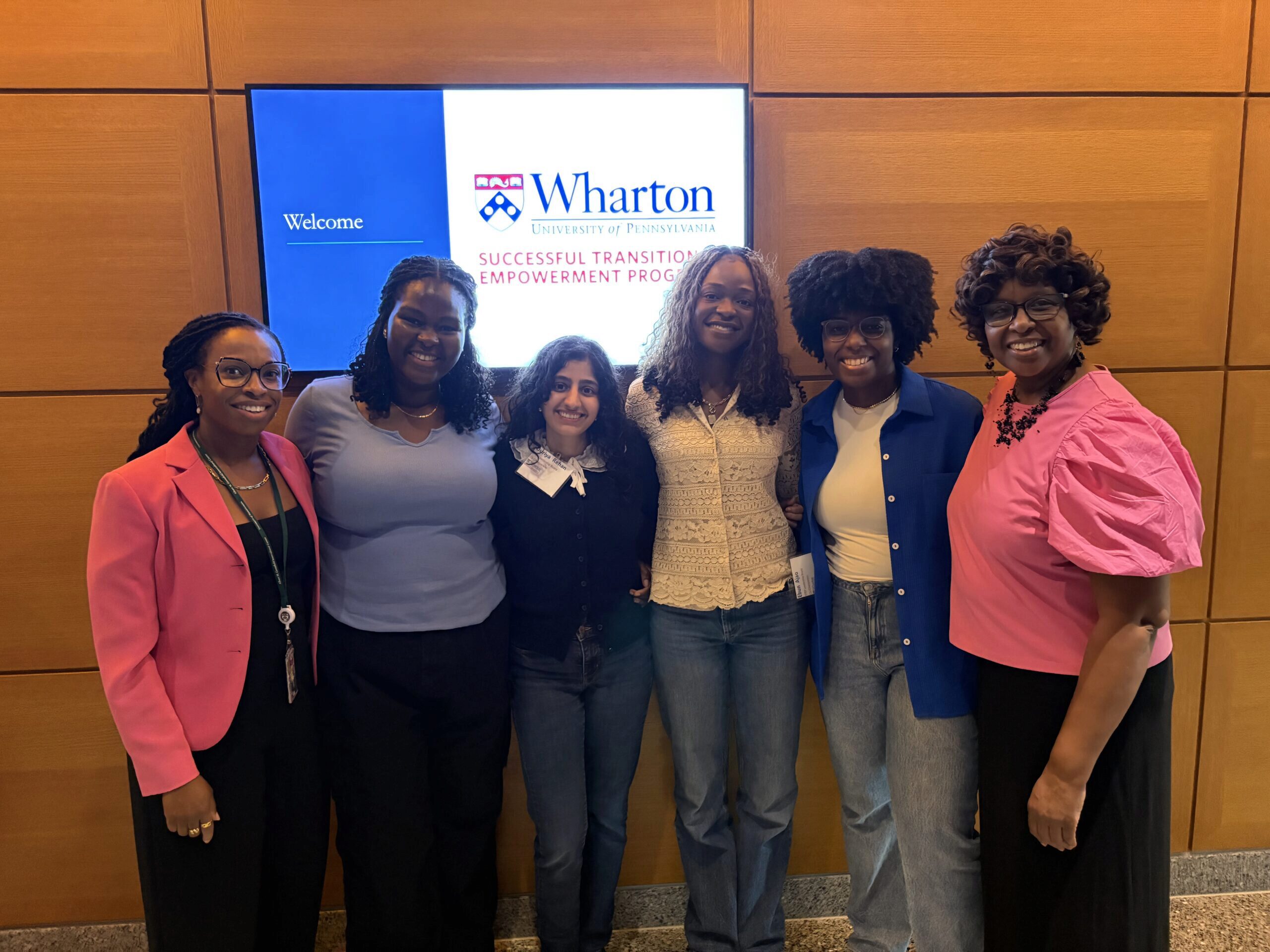IMG_7114 A group of people standing in front of a Wharton University of Pennsylvania sign, dressed in business casual attire, smiling at the camera.