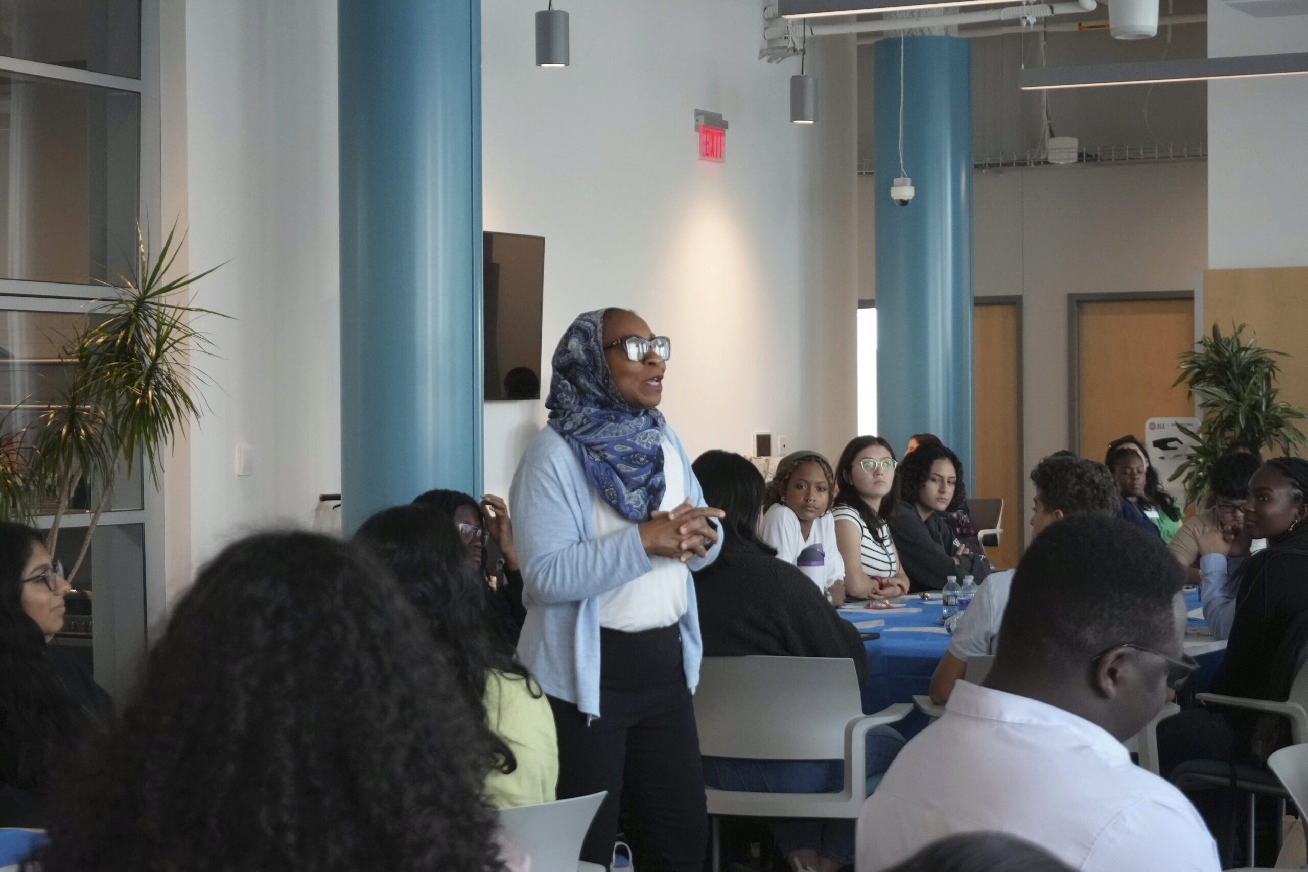 STEP Day 3-120 A person wearing a hijab and sunglasses is speaking to a diverse group seated at tables in a conference or workshop setting.