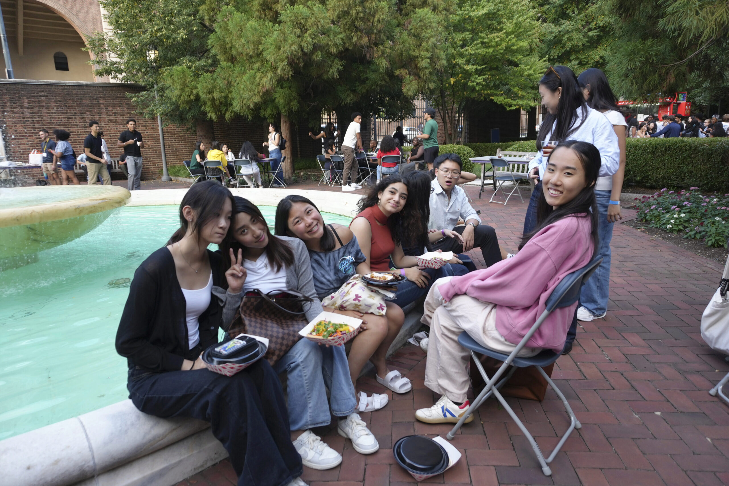 STEP Day 3-152 A group of young people gathered around a fountain in an outdoor setting, some sitting on the edge and others nearby on chairs, with trees and brick pathways in the background.