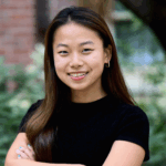 Headshot of a person with long hair, smiling, and wearing a black shirt, standing outdoors with greenery in the background.