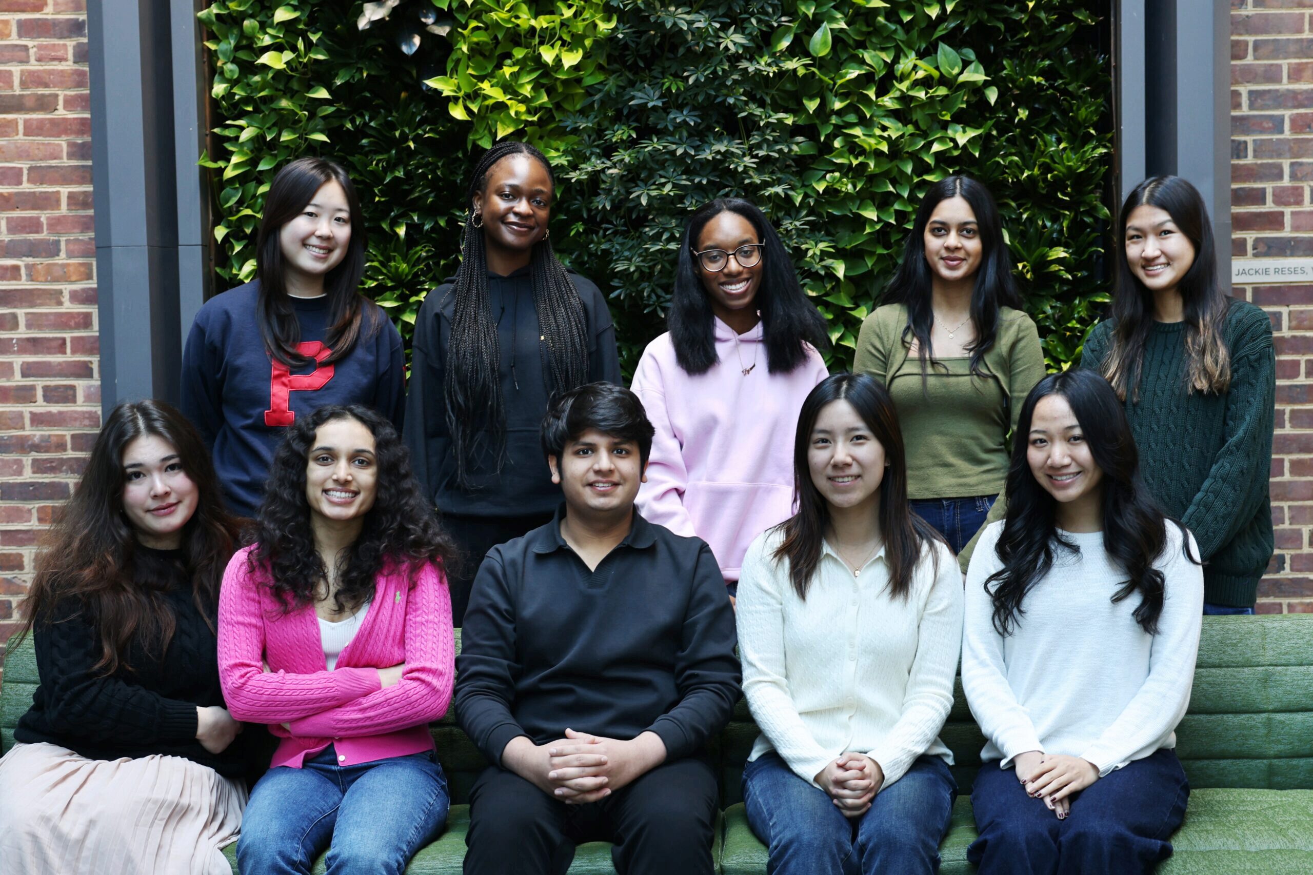 Ten students pose in two rows before a lush green wall and brick courtyard, smiling at the camera.